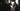 Woman painting at a easel in a dimly lit room with other people in the background.